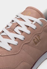 Gros plan sur une basket rose poudré avec des lacets blancs et le logo "GAP" doré sur le côté et la languette.