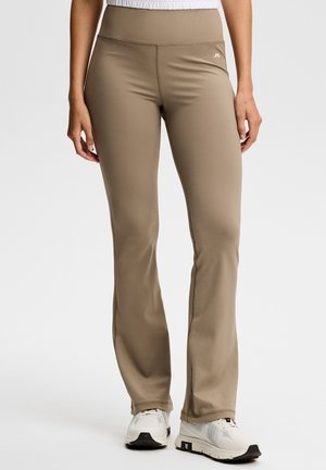 Donna che indossa pantaloni beige a vita alta e aderenti e scarpe da ginnastica bianche, in piedi davanti a uno sfondo chiaro e semplice.