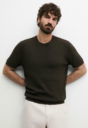 Uomo con capelli ricci e barba che indossa un maglione corto a maniche corte scuro e pantaloni chiari, in piedi con le mani dietro la schiena.