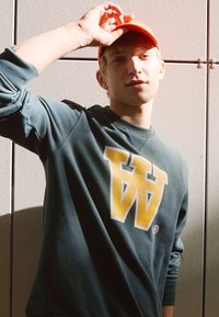 Grüner Sweatshirt mit einem großen gelben "W"-Logo, getragen mit einer orangen Mütze. Die Textur wirkt weich und der Schnitt ist lässig. Der Hintergrund ist neutral.