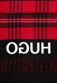 Tissu à carreaux rouge et noir avec franges noires et lettres majuscules blanches « HUGO » imprimées en miroir sur fond noir.