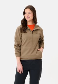 Brauner, leichter Fleece-Pullover mit Reißverschluss und hohem Kragen, langen Ärmeln und einer Tasche. Trägt man über einem orangefarbenen Shirt, kombiniert mit schwarzen Hosen.