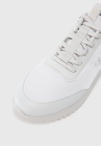 Λευκό αθλητικό παπούτσι με λείο υλικό άνω τμήματος με ριγέ σχέδιο, επίπεδες κορδόνες και μαλακή σόλα. Ολογραφικό λογότυπο ορατό σ�τη μεριά.