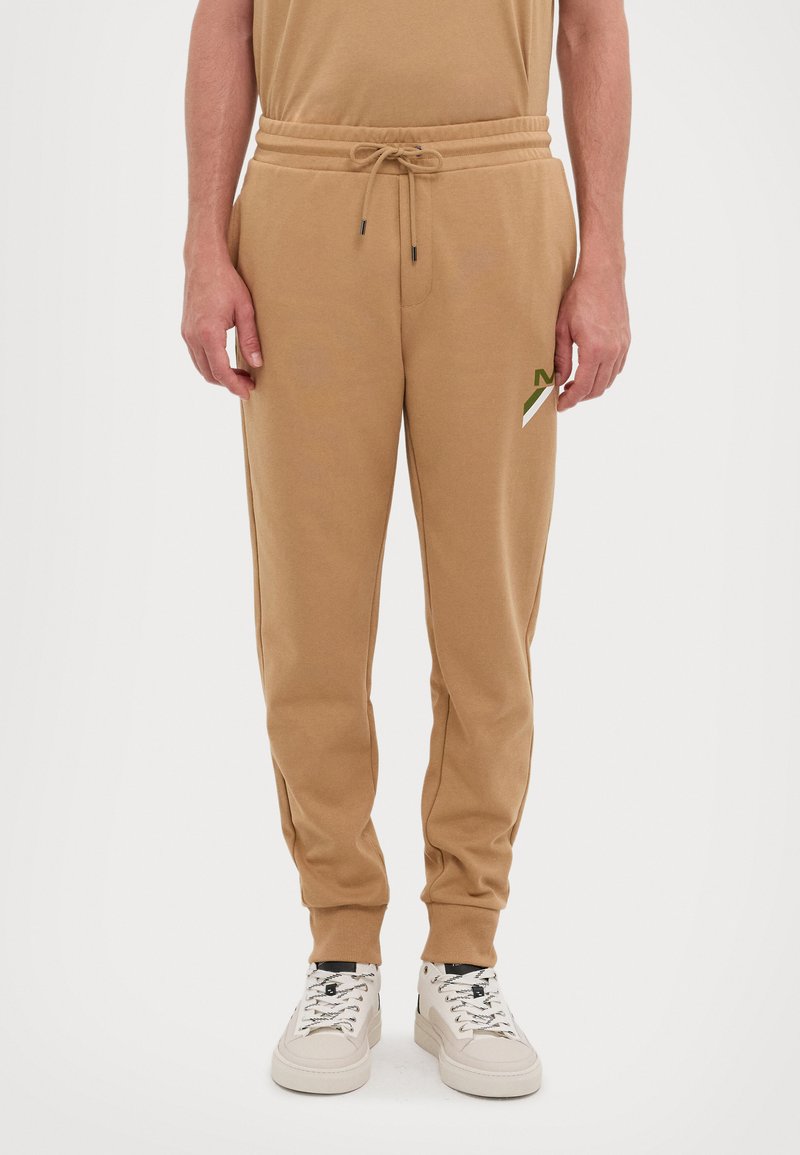 Pantalones jogger en color marrón claro con cintura elástica, cierre con cordón, bolsillos laterales y un pequeño logo en verde y blanco en el muslo izquierdo.