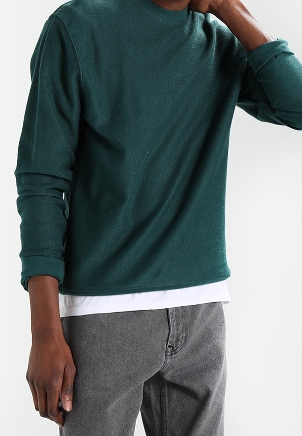 Homme portant une chemise à manches longues vert foncé sur un sous-vêtement blanc, associée à un jean gris, main touchant le cou.