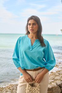 Femme en blouse turquoise et pantalon beige tenant un sac décoré de coquillages, debout sur un rivage rocheux avec l'océan en arrière-plan.