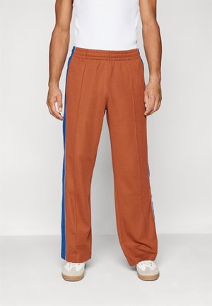 Homme portant un pantalon large couleur rouille avec des bandes latérales bleues, associé à des baskets blanches et un débardeur blanc.