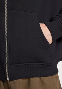 Black sweatshirt with a zippered front, ribbed hem, and pockets. Made of textured fabric, featuring a smooth zipper and fitted cuffs.