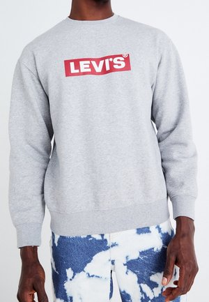 Männliches Model trägt ein hellgraues Levi’s Sweatshirt kombiniert mit blau-weißen Batik-Denim-Jeans und steht vor einem einfarbigen Hintergrund.