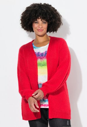 Cardigan rouge en tricot avec manches longues, design ouvert à l'avant et tissu texturé. Porté par-dessus une chemise tie-dye multicolore et un jean noir.