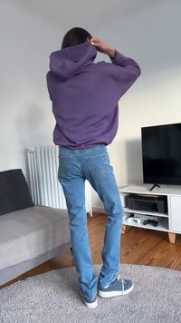 Camisola com capuz roxa com bolso canguru, jeans de ganga azul e ténis com padrão. Cena apresenta uma sala de estar minimalista com paredes brancas.