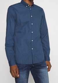 Camicia blu navy con bottoni e piccolo motivo geometrico ripetuto. Presenta un colletto classico e bottoni bianchi. Indossata con jeans in denim.