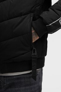 Hand in der Tasche einer schwarzen gesteppte Jacke mit Bündchen, Reißverschluss und angebrachtem schwarzen Stoff-Ziehschlaufe mit aufgedruckten Zahlen, getragen über grauen Jeans.