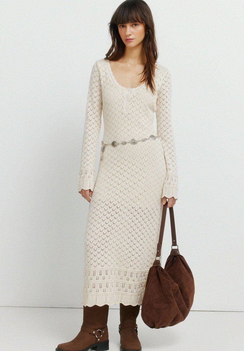 Žena ve smetanově krémových pletených šatech s dlouhým rukávem se stříbrným páskem, hnědými semišovými kozačkami a s velkou hnědou semišovou kabelkou v ruce.