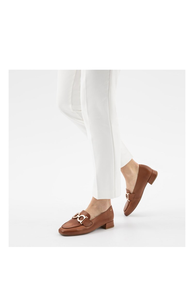 Mocassins en cuir marron avec détail chaîne dorée portés avec un pantalon blanc raccourci sur un fond blanc uni.