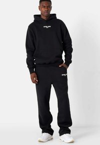 Schwarzer Hoodie mit Fronttasche und weißem Text, kombiniert mit schwarzen Jogginghosen mit Seitentaschen. Die Schuhe sind weiß mit hellbraunen Akzenten.