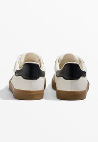 Weiße Sneakers mit einem schwarzen Fersenelement, ausgestattet mit einer strukturierten Gummisohle und Stoffsenkeln. Das Design ist tief ausgeschnitten mit abgerundeten Kanten.