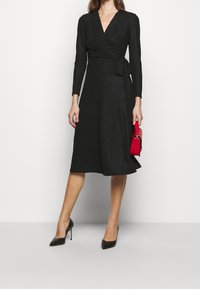 Nainen, joka käyttää mustaa pitkähihasta kietaisumekkoa, mustia korkokenkiä ja pitää kädessään pientä punaista käsilaukkua vaalean valkoista taustaa vasten.