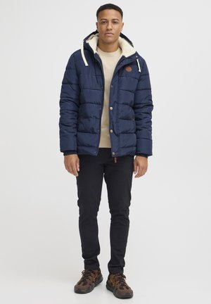 Marineblå pufferjakke med isoleret design, krème-foring i hætten og brune knapaccentuer, parret med sorte bukser og brune mønstrede sneakers.