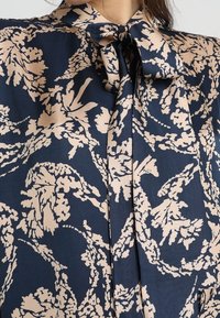 Marinblus med blommönster i krämfärg, med halsband och knappstängning. Tillverkad av ett lätt och slätt tyg.