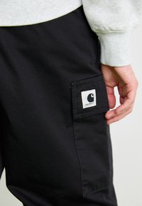 Zwarte cargobroek met een zijzak voorzien van een wit Carhartt-logo. Het materiaal lijkt duurzaam met een eenvoudige textuur en een nauwsluitende pasvorm.