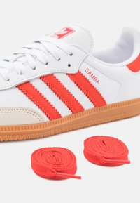 Vita Adidas Samba-sneaker med röda accenter, tre röda ränder och en beige tåhuvud; inkluderar två rullade röda skosnören.