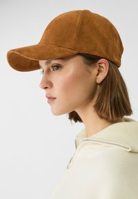 Casquette de baseball en daim marron avec une visière courbée, portée à un angle, dotée d'un design cousu. L'arrière-plan est neutre et minimaliste.