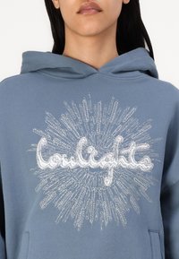 Sweatshirt azul com uma imagem na frente apresentando a palavra "Coulights" em letra cursiva branca, rodeada por um design de raios de sol. Feito de um tecido macio.