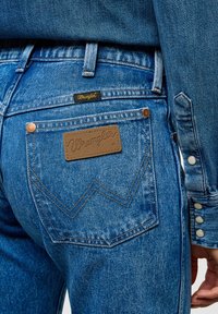 Mėlyni džinsai su odine Wrangler etikete ant gale esančio kišenės, vario varžtais ir išskirtiniu zigzago siuvinėjimu ant kišenių.