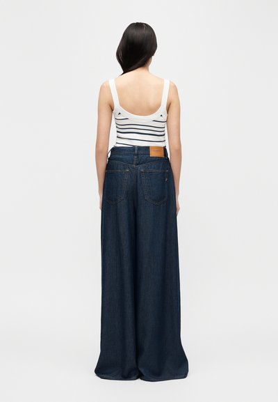 Woman with long dark hair wearing a white striped tank top and high-waisted, wide-leg dark blue jeans, standing facing away.