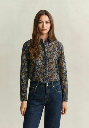 Jeune femme aux longs cheveux bruns portant une chemise boutonnée à motif floral et un jean bleu foncé taille haute sur fond uni.