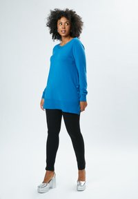 Blaues langärmeliges Sweatshirt mit rundem Ausschnitt und seitlichen Knöpfen, kombiniert mit schwarzen Leggings und silbernen Plateauschuhen, auf einem Modell.