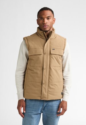 Mann trägt eine beige wattierte Weste über einem cremefarbenen langärmeligen Shirt und blauen Jeans und steht vor einem schlichten grauen Hintergrund.