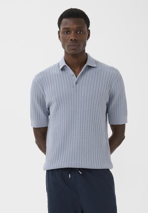 Mann mit kurzen lockigen Haaren steht frontal und trägt ein hellblaues kurzärmliges Kabelstrick-Poloshirt und dunkelblaue Hose mit Kordelzug.