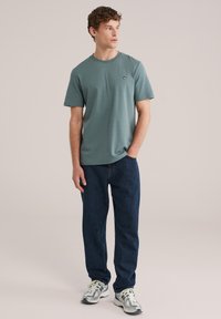 Jonge man staand, draagt een gedempt groene korte mouw T-shirt, donkerblauwe jeans en witte sneakers met groene accenten, kijkt zijwaarts.