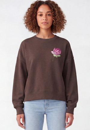 Femme aux cheveux bouclés portant un sweat-shirt marron avec un motif de fleur rose sur le côté gauche de la poitrine et un jean bleu clair sur fond blanc.