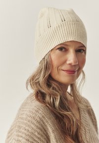 Krémová pletená čiapka s ryhovanou textúrou a malým horným prehnutím. Mäkká látka s vertikálnymi vzormi. Nosí ju osoba s kučeravými vlasmi.