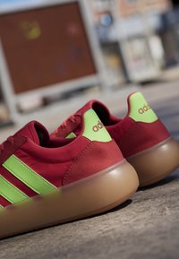 Pair of red and neon green Adidas sneakers with gum soles placed on outdoor pavement in soft sunlight.