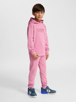 Dreng står iført en pink hættetrøje og matchende joggingbukser med blå og grå sneakers, foran en ensfarvet hvid baggrund.