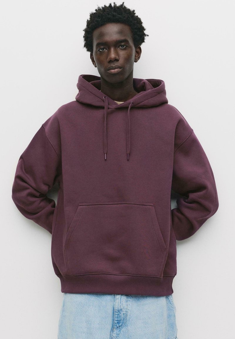 Felpa bordeaux con cappuccio e coulisse, grande tasca frontale e polsini a costine. Tessuto morbido e vestibilità rilassata, abbinata a jeans chiari.