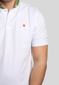 Polo bianco con colletto corto, con una fodera a motivo verde e un logo arancione sul petto. Tessuto morbido e leggero.