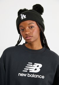 Fekete kötött sapka pom-pomokkal, fehér "LA" logóval. A modellen sötét pulcsi van, amelyen nagy fehér "new balance" logó látható.