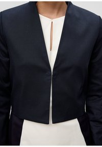 Diep marineblauwe cropped blazer met een strak ontwerp, voorzien van een minimalistische sluiting aan de voorkant en een sjawl-kraag, gecombineerd met een lichtcrème top eronder.