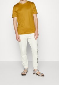 Senapsgul kortärmad skjorta, vita joggers med sidostripes, och grå sandaler med justerbara remmar. Slätt tyg, figursydd design.