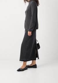 Femme portant un pull à col roulé côtelé gris foncé, une jupe en denim noire longueur cheville avec ourlet effiloché, des ballerines noires pointues, tenant un petit sac noir.