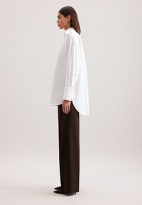 Camicia bianca oversize con colletto alto e polsini abbottonati, abbinata a pantaloni marroni a gamba dritta e scarpe con punta.