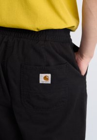 Pantalones negros con cintura elástica, que cuentan con un bolsillo trasero con una etiqueta cuadrada que incluye un logo amarillo. Material de algodón suave.