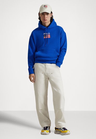 Blå hettegenser med "POLO" og flaggemblem, kombinert med kremfargede bukser og flerfargede joggesko. Bomullsmateriale, avslappet passform, hette design.