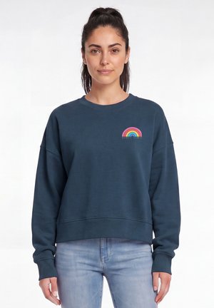 Junge Frau mit dunklen Haaren im Pferdeschwanz trägt einen dunkelblauen Pullover mit einem kleinen bunten Regenbogen und dem Text "Women Power" auf der Brust.