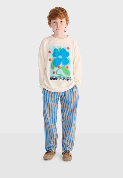 Garçon aux cheveux roux portant un haut à manches longues crème avec une fleur bleue et du texte, un pantalon rayé bleu et beige, ainsi que des chaussures marron, debout les mains dans les poches.